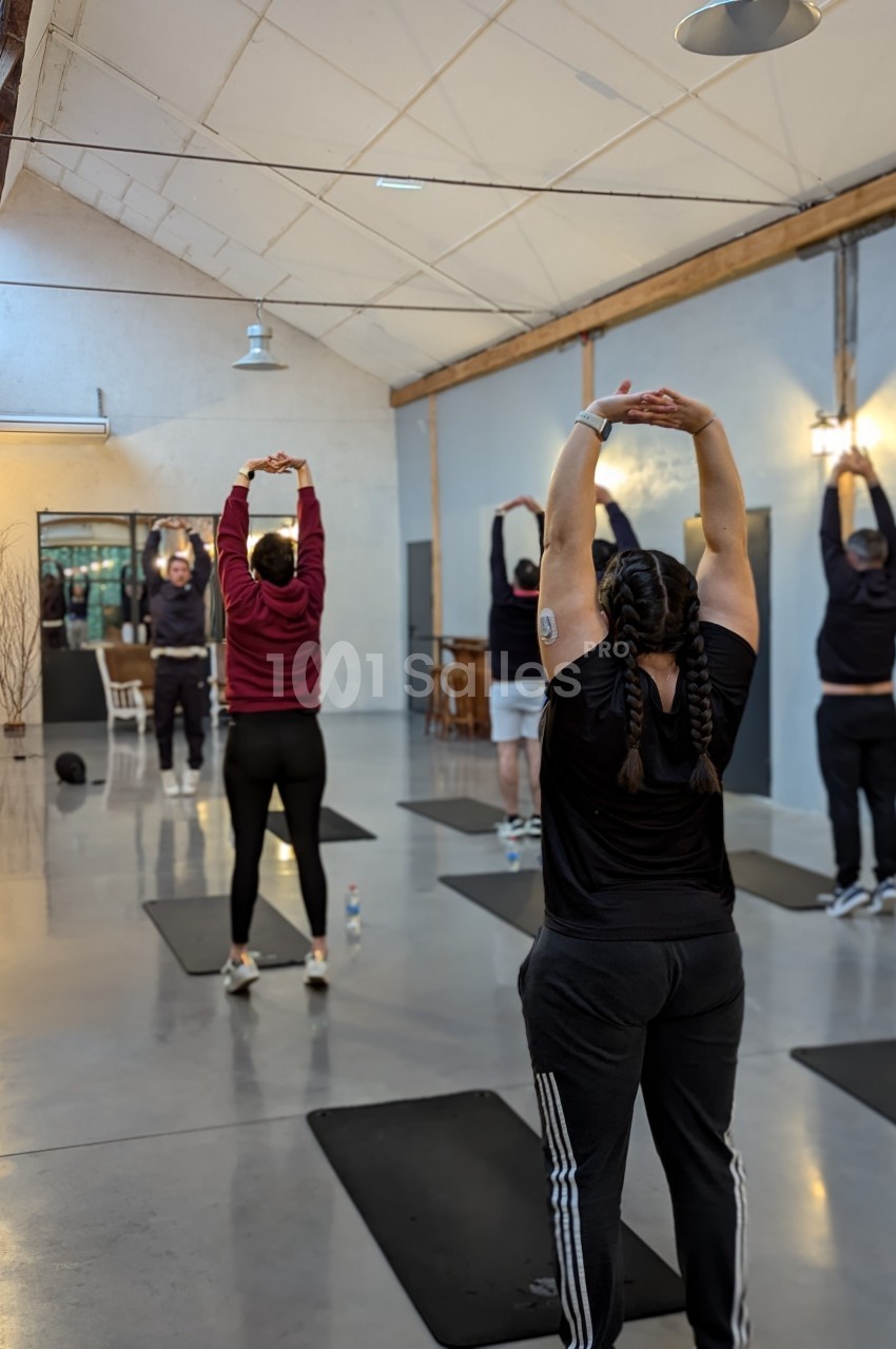 Personnes s'étirant les bras levés dans une salle de sport lumineuse, debout sur des tapis de yoga.