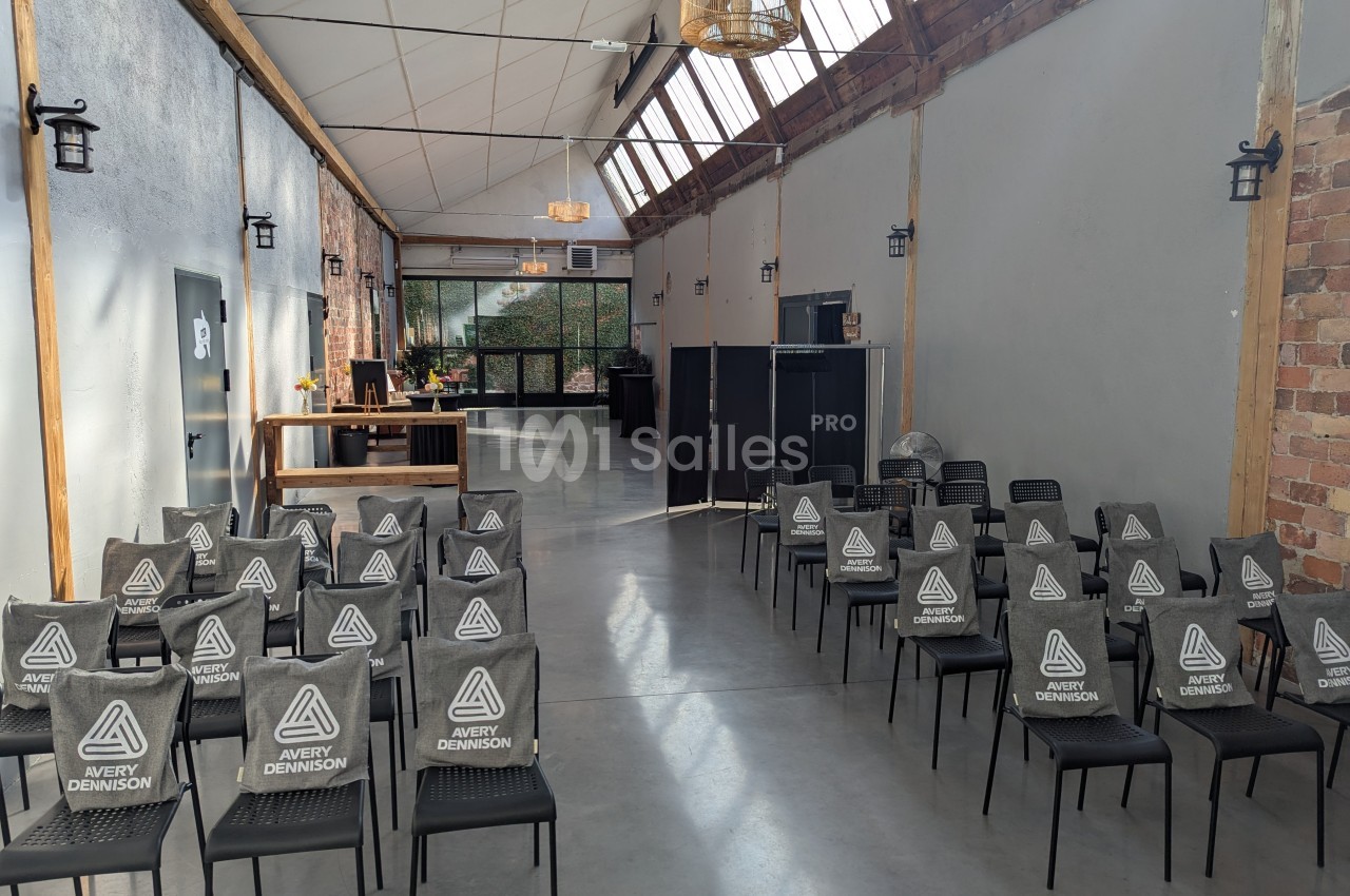 Salle lumineuse avec des rangées de chaises noires alignées, un comptoir en bois et des murs en briques apparentes.