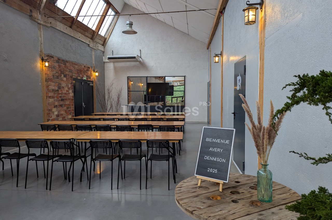 Salle lumineuse aménagée avec des tables en bois, des chaises noires et une décoration végétale, prête pour un événement.