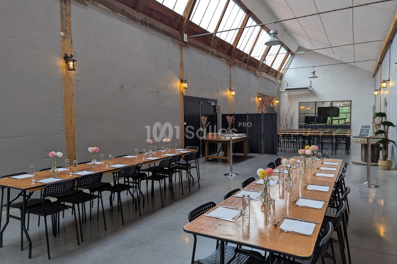 Salle lumineuse avec de grandes tables en bois dressées, chaises noires, décor minimaliste et verrière en hauteur.