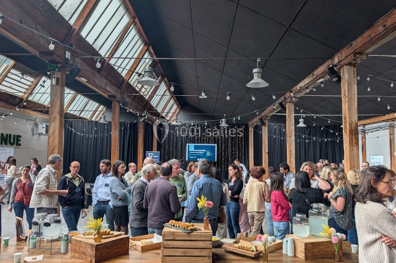 Groupe de personnes échangeant dans une grande salle avec toit vitré, tables en bois et buffet au premier plan.