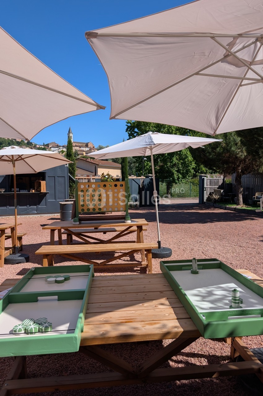 Tables en bois avec jeux de palets, parasols blancs et décor extérieur ensoleillé avec arbres et cabane en arrière-plan.