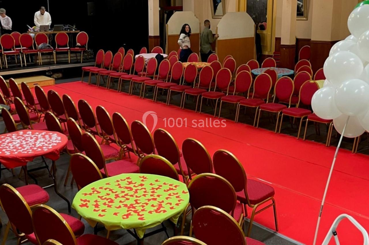Salle décorée avec une scène, des chaises alignées autour d'un tapis rouge et des tables ornées de nappes colorées.