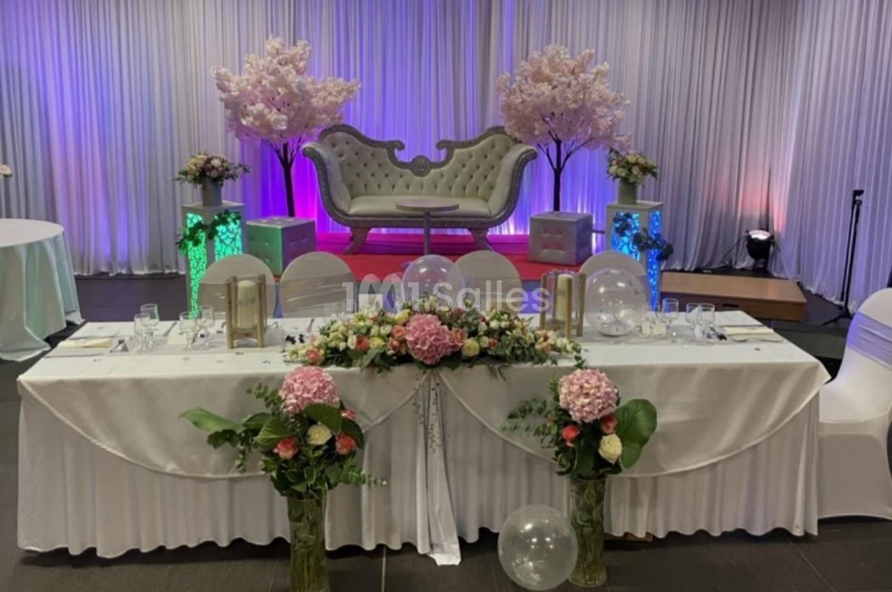 Table décorée avec des fleurs et des ballons devant un canapé élégant, dans une salle ornée de rideaux et éclairages colorés.
