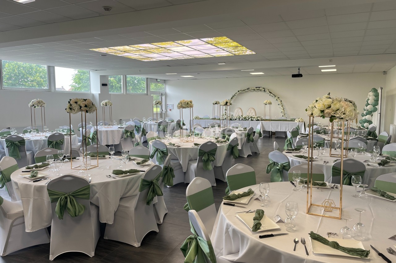 Salle de réception décorée avec des tables rondes, nappes blanches, chaises ornées de nœuds verts et centres de table…