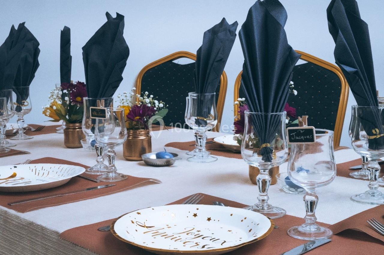 Table dressée avec vaisselle élégante, serviettes noires pliées, fleurs décoratives et marque-places sur fond clair.