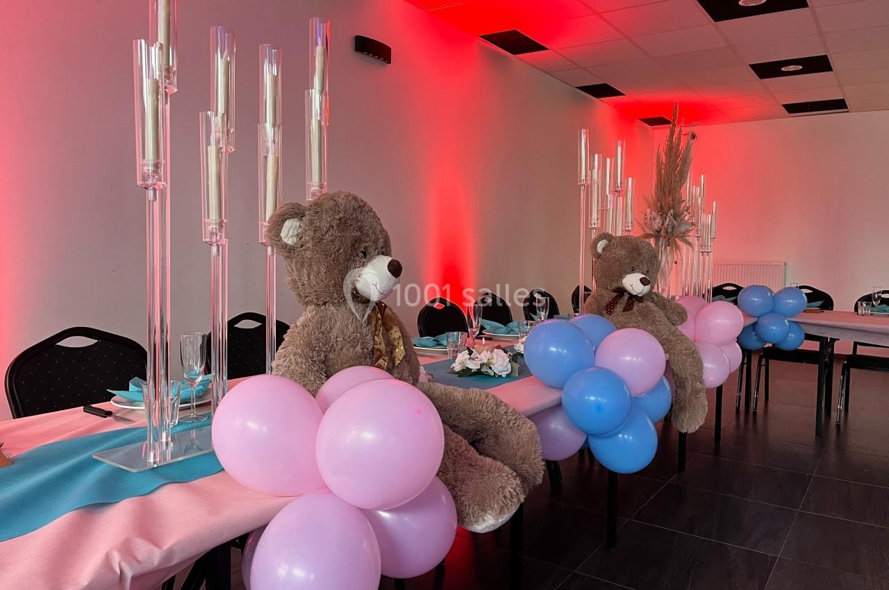 Deux ours en peluche décorent une table ornée de ballons roses et bleus dans une salle éclairée par des lumières rouges.