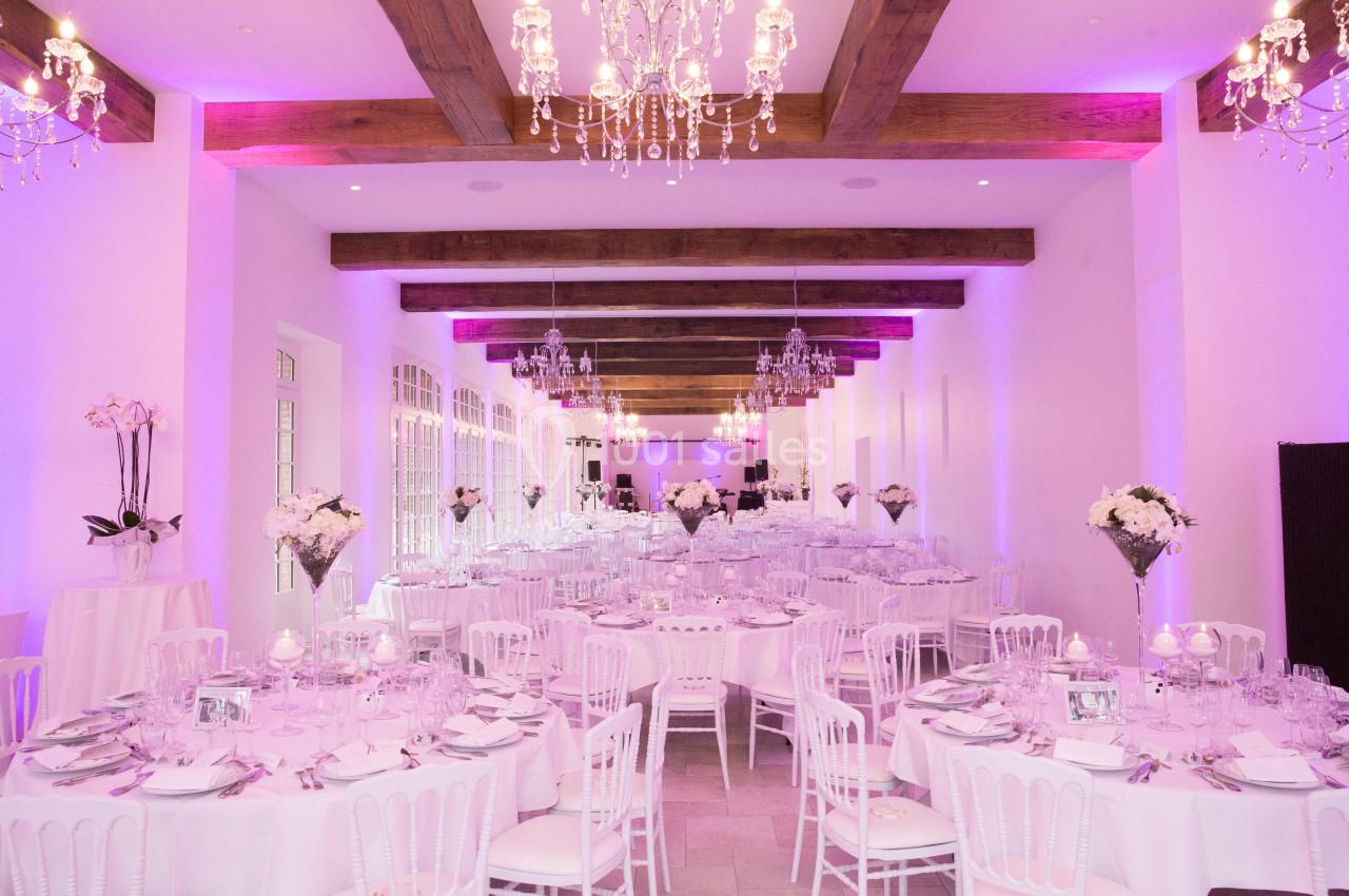 Salle de réception élégante avec tables rondes décorées, chaises blanches et éclairage rose sous des lustres.