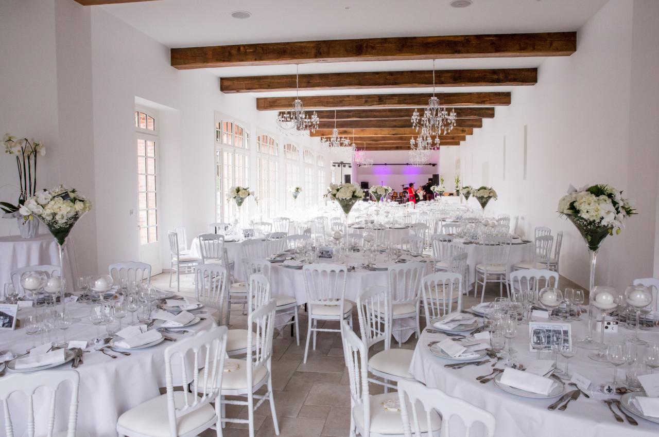 Salle de réception lumineuse avec tables décorées de nappes blanches, chaises assorties et arrangements floraux élégants.