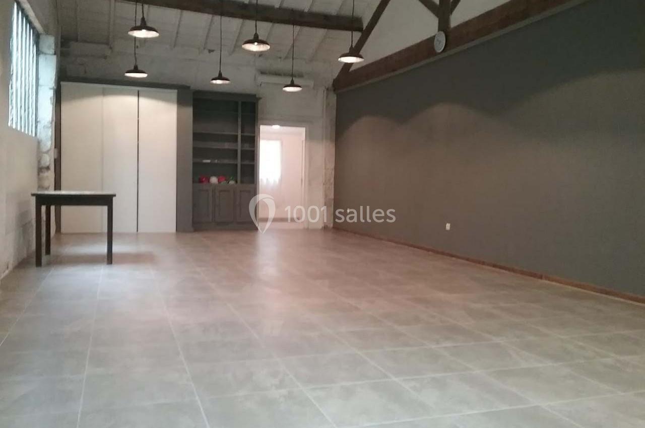 Salle vide avec sol en carrelage, murs gris et blancs, poutres apparentes au plafond, éclairée par des suspensions.
