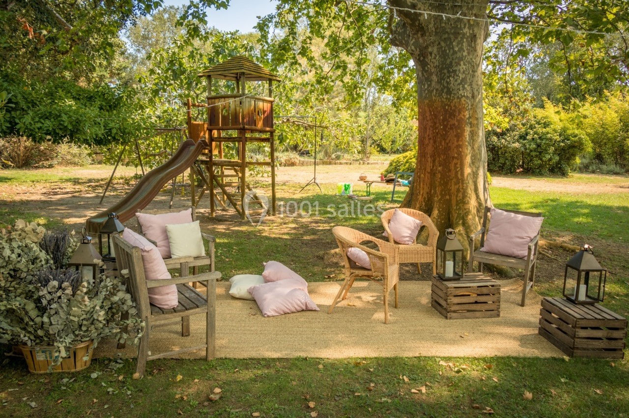 Espace extérieur avec fauteuils, coussins, lanternes et une aire de jeux en bois sous un grand arbre.