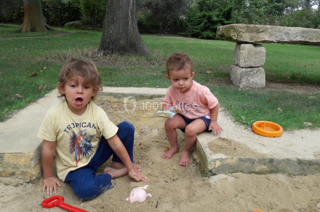 Deux jeunes enfants jouent dans un bac à sable, entourés de verdure et de jouets en plastique.