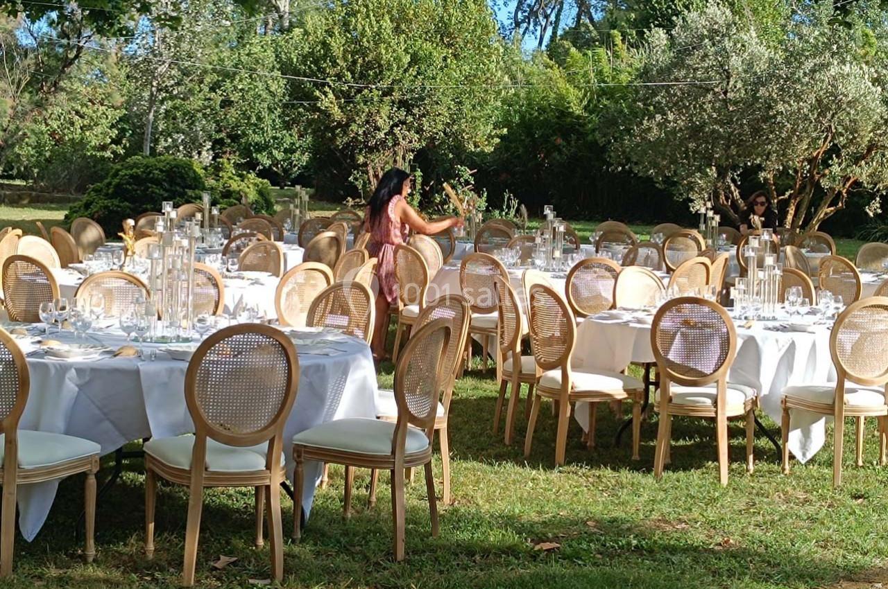 Tables rondes dressées avec nappes blanches et chaises en bois dans un jardin, préparées pour un événement en plein air.