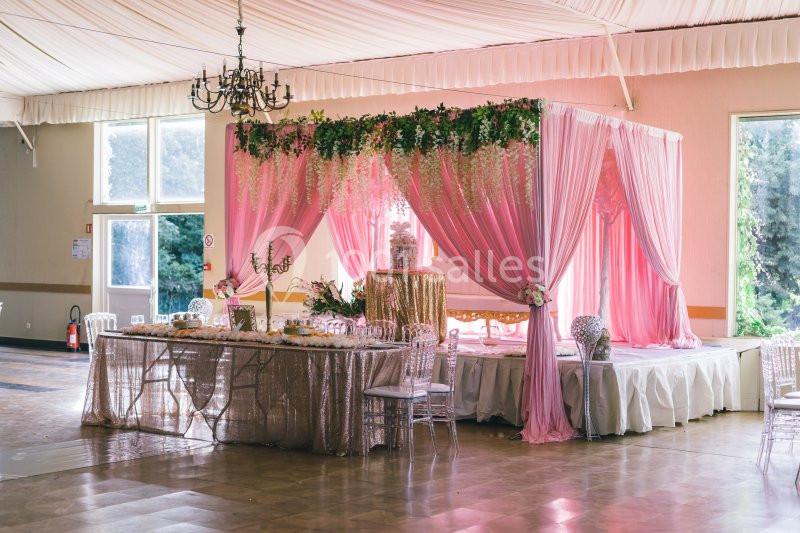 Salle de réception décorée avec des rideaux roses, une table ornée et un espace surélevé sous un dais floral.