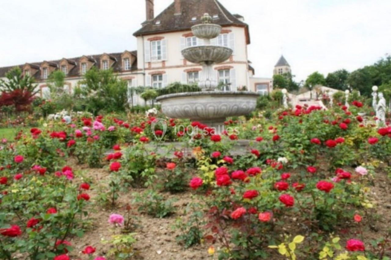 Jardin fleuri avec des rosiers rouges et une fontaine en pierre devant une grande maison à toit en pente.
