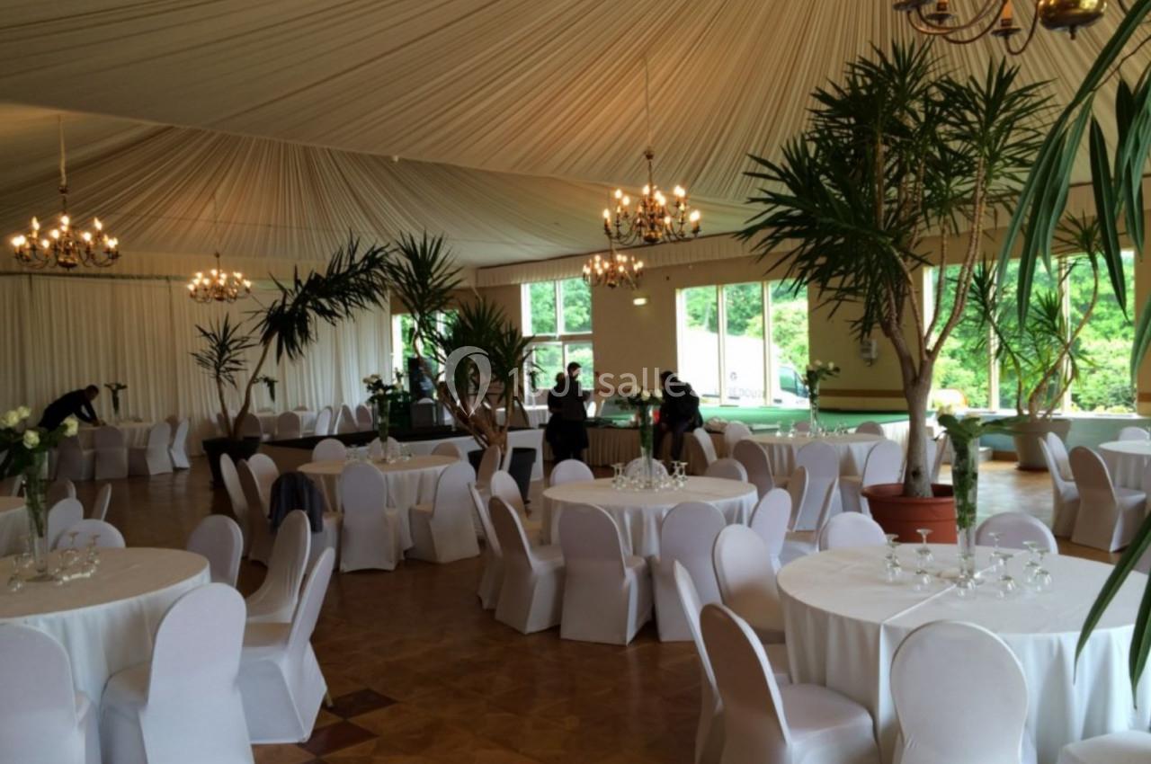 Salle de réception élégante avec tables rondes dressées, chaises blanches et grandes plantes décoratives.