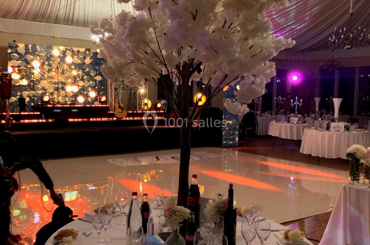 Table dressée avec nappes blanches, couverts, bouteilles et pain, décorée d'un arbre fleuri sous une tente éclairée.