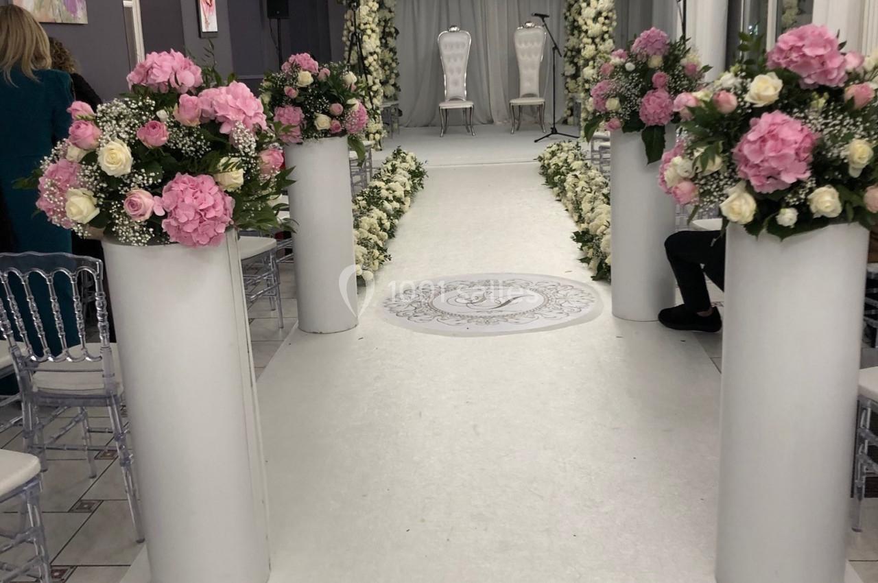 Allée décorée de fleurs blanches et roses menant à une arche florale, dans une salle éclairée par des lustres.