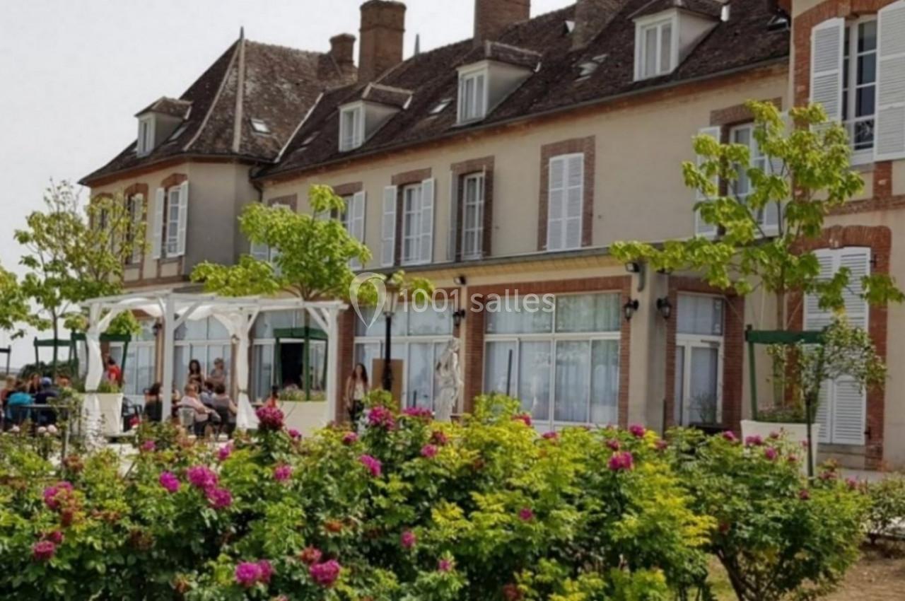 Façade d'un grand bâtiment ancien avec des volets blancs, entouré de rosiers et d'une terrasse occupée par des personnes.
