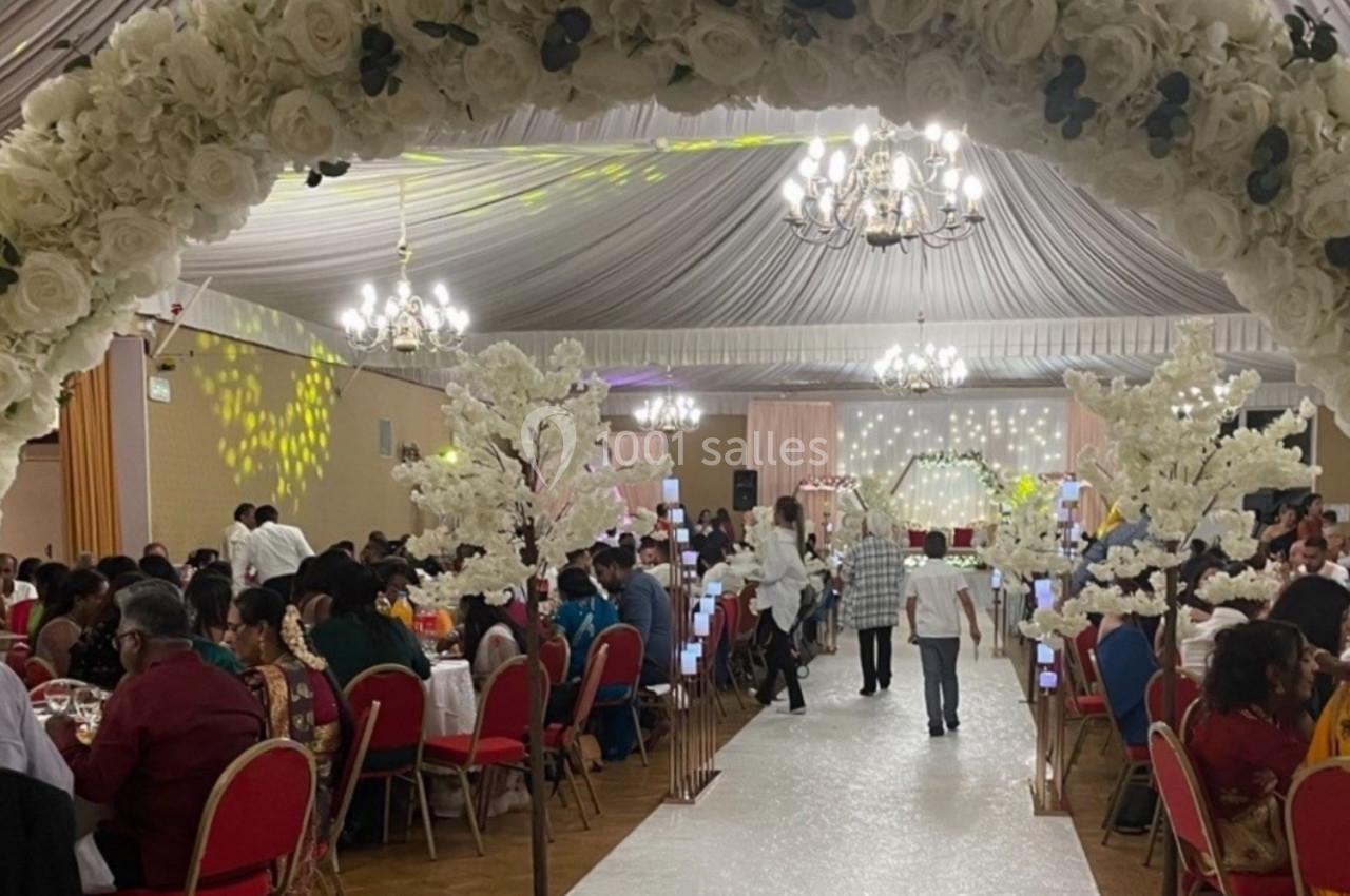 Salle de réception décorée pour un événement, avec une arche de fleurs blanches et des invités assis sur des chaises rouges.