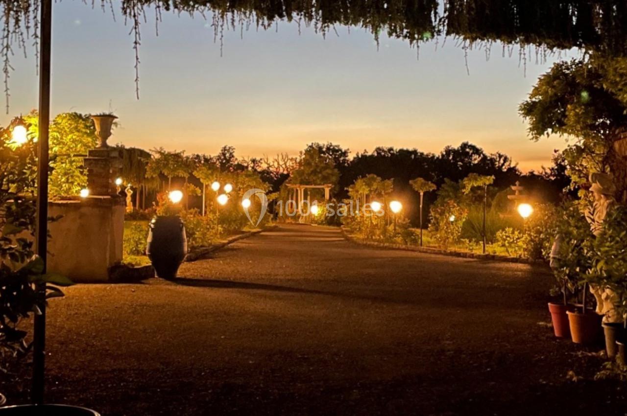 Allée de jardin éclairée par des lampes, bordée de plantes en pot, au crépuscule avec un ciel orangé.