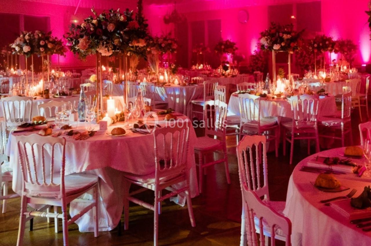 Salle de réception décorée avec des tables rondes, nappes blanches, chandeliers et éclairage rose tamisé.