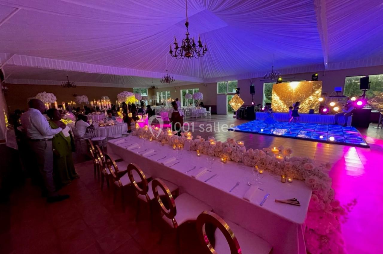 Salle de réception décorée avec des tables ornées de fleurs et bougies, piste de danse illuminée en arrière-plan.