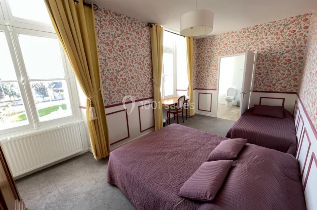 Chambre lumineuse avec deux lits, murs tapissés de motifs floraux, fenêtres avec rideaux jaunes et bureau en bois.