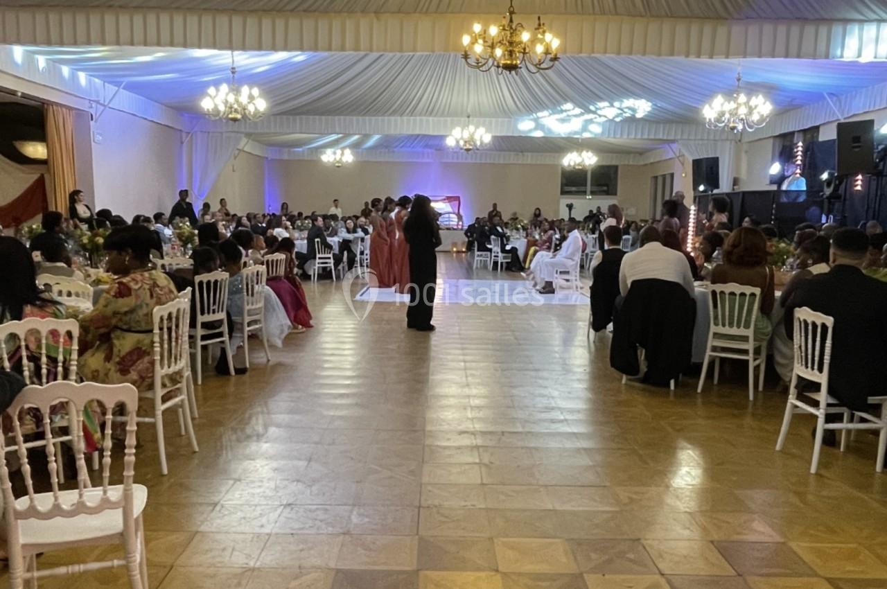 Salle de réception éclairée avec des invités assis à des tables et des personnes dansant au centre.