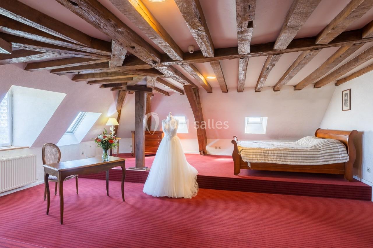 Chambre mansardée avec poutres apparentes, lit en bois, robe de mariée sur mannequin et table avec bouquet de fleurs.