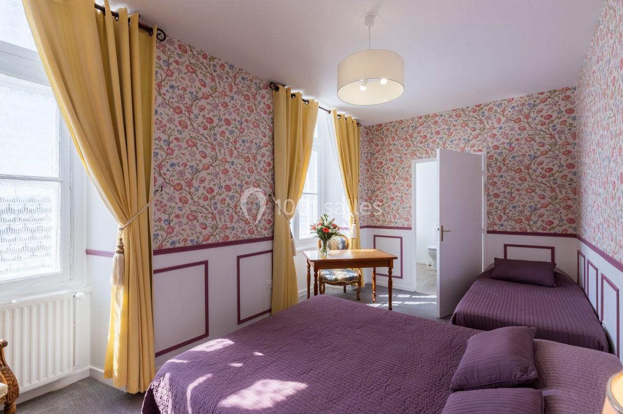 Chambre lumineuse avec deux lits, murs à motifs floraux, rideaux jaunes et une table avec un vase de fleurs.