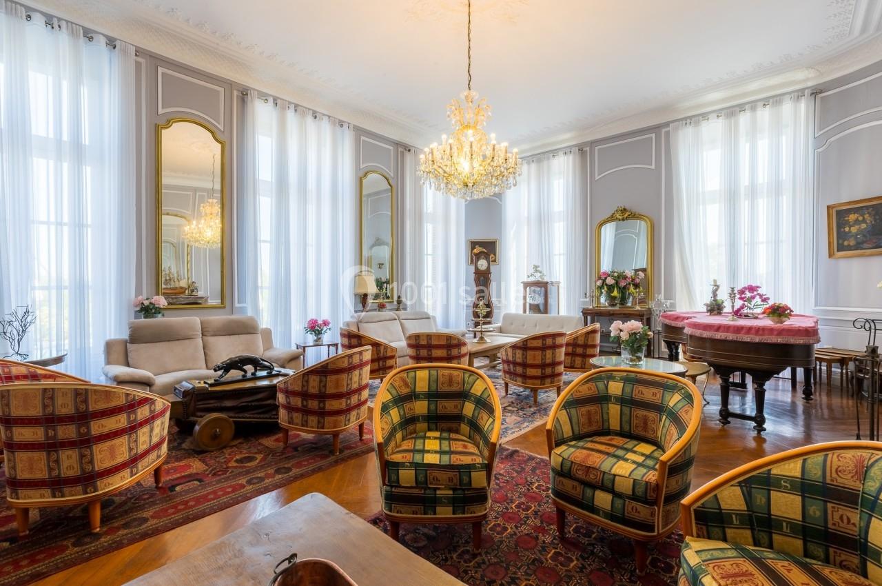 Salon élégant avec fauteuils colorés, lustre central, miroirs dorés, piano et grandes fenêtres aux rideaux blancs.