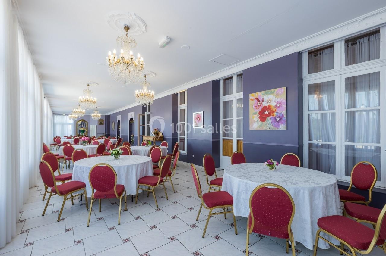 Salle lumineuse avec des tables rondes dressées de nappes blanches, chaises rouges et lustres suspendus.
