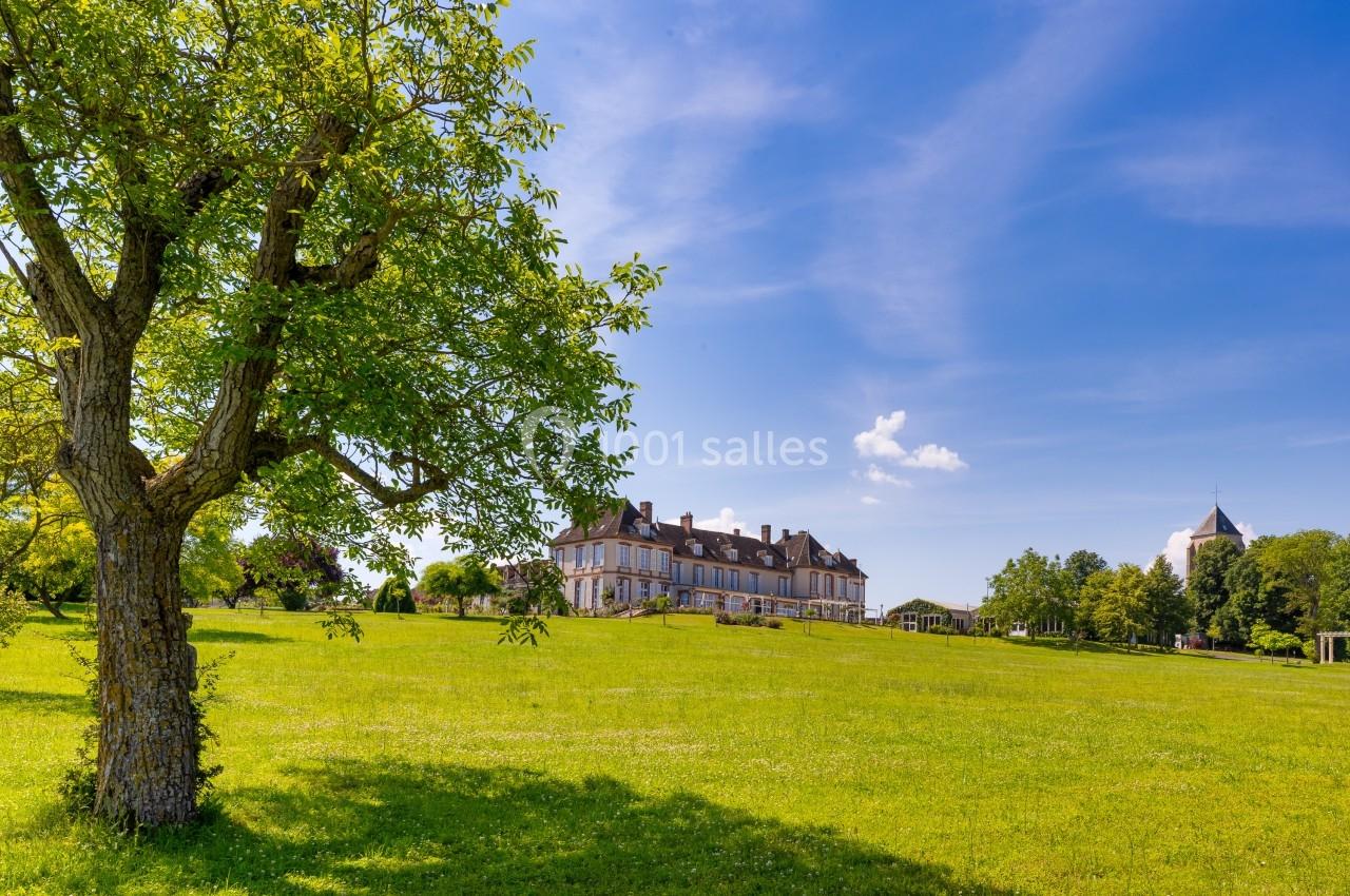 Grand bâtiment entouré d'une vaste pelouse verte avec un arbre au premier plan, sous un ciel bleu dégagé.