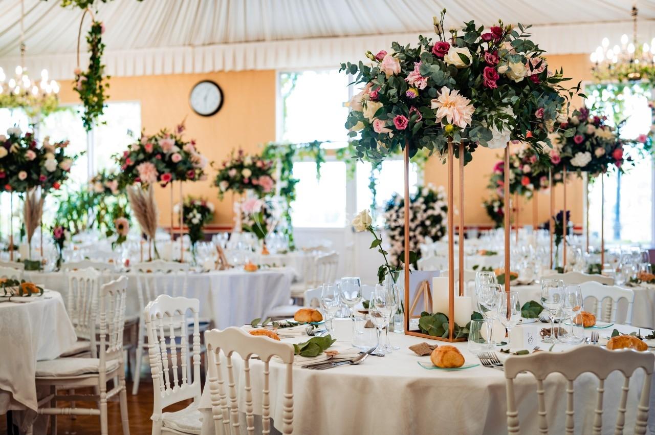 Salle de réception décorée avec des tables rondes, nappes blanches, centres de table fleuris et chaises blanches.