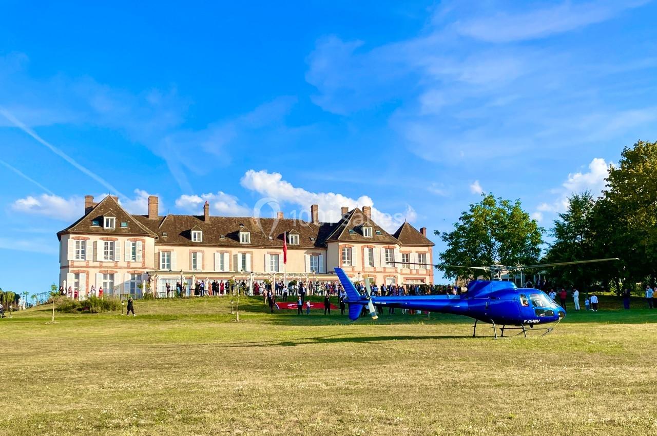 Hélicoptère bleu posé sur une pelouse devant un grand bâtiment historique sous un ciel dégagé.