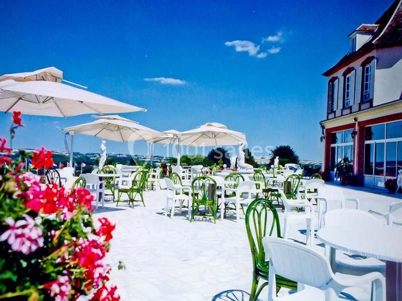 Terrasse ensoleillée avec tables, chaises blanches et parasols, entourée de fleurs et offrant une vue dégagée sur la…