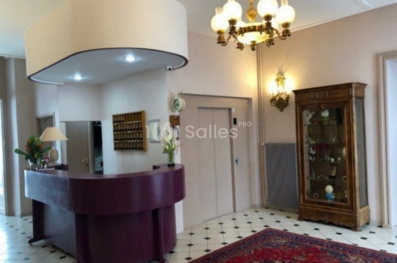 Accueil d'un hôtel avec comptoir violet, lustre ancien, vitrine en bois et tapis rouge sur un sol carrelé.