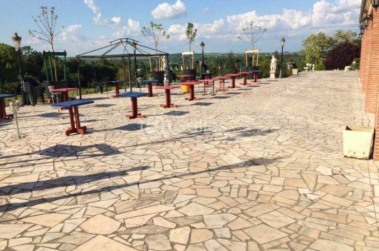 Terrasse pavée avec des tables colorées espacées, entourée de verdure et offrant une vue dégagée sur l'horizon.