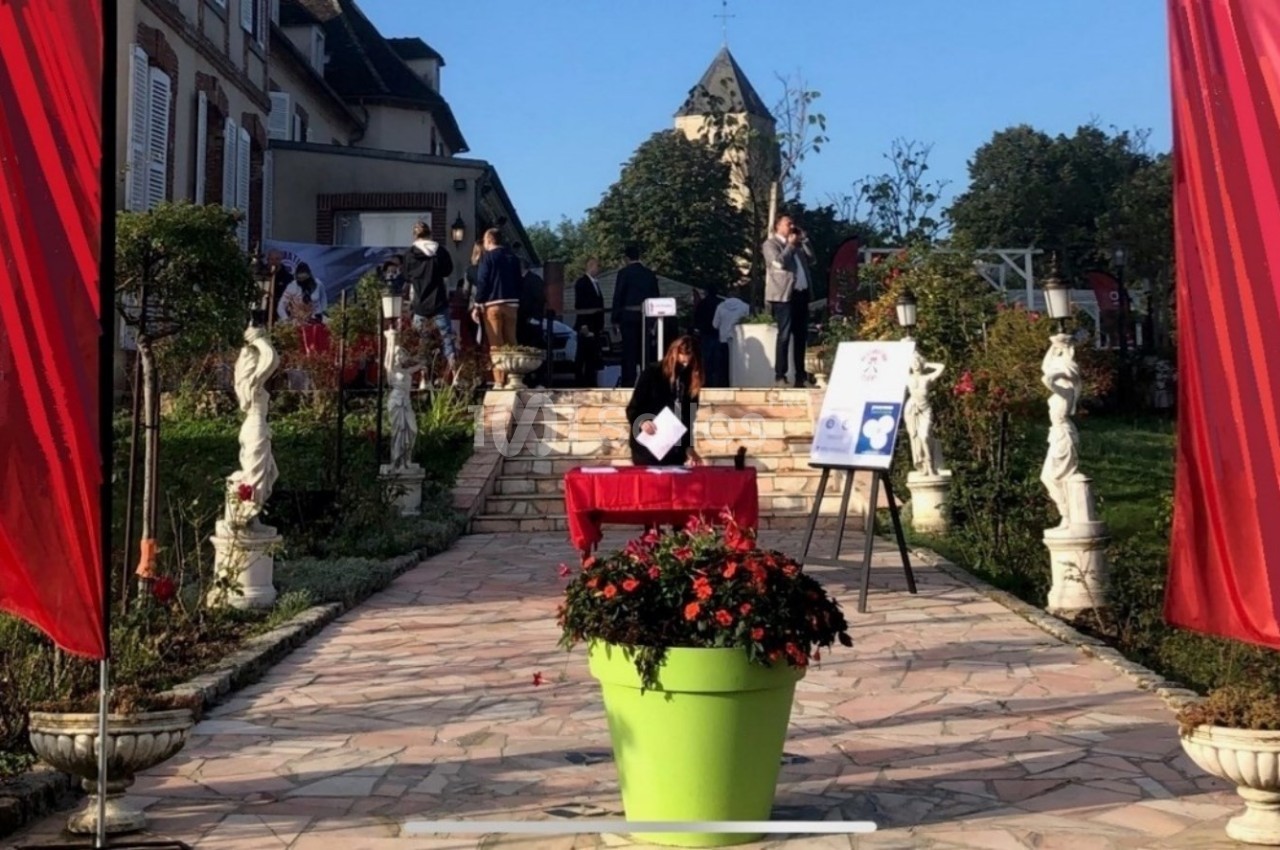 Allée pavée bordée de statues et de plantes, menant à une table rouge devant un bâtiment avec un clocher en arrière-plan.