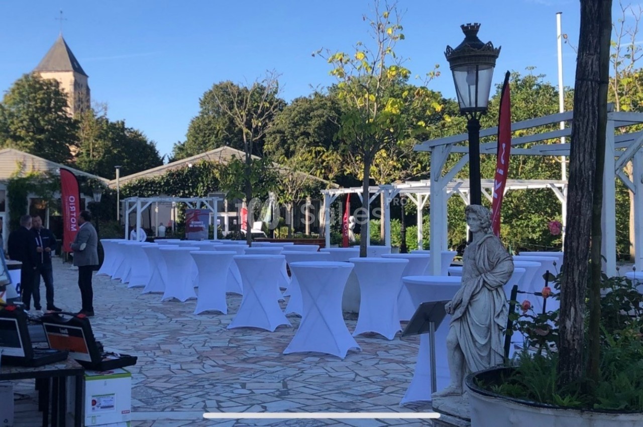 Terrasse extérieure avec tables hautes dressées sous des pergolas blanches, entourée de verdure et d'une statue.