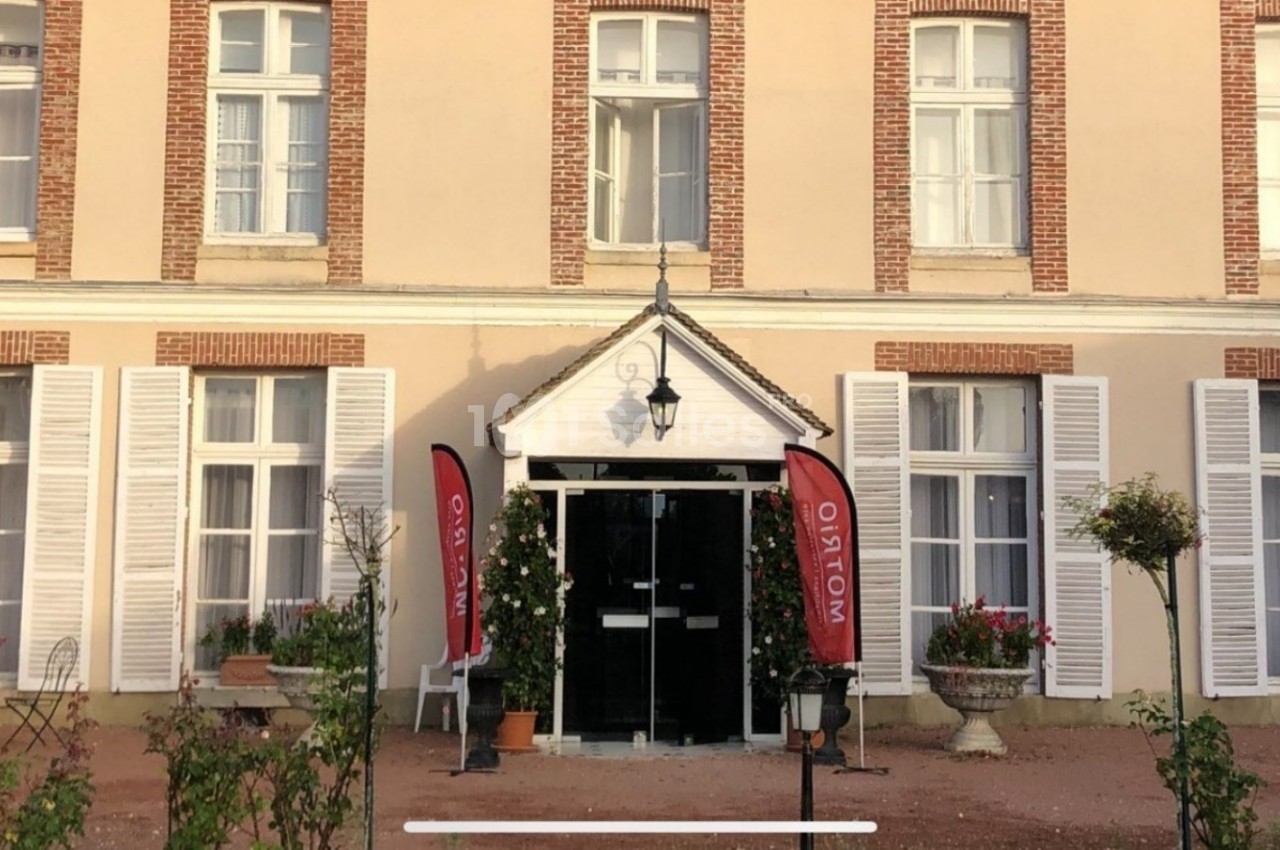 Façade d'un bâtiment avec des volets blancs, une entrée centrale et deux drapeaux rouges près de la porte.