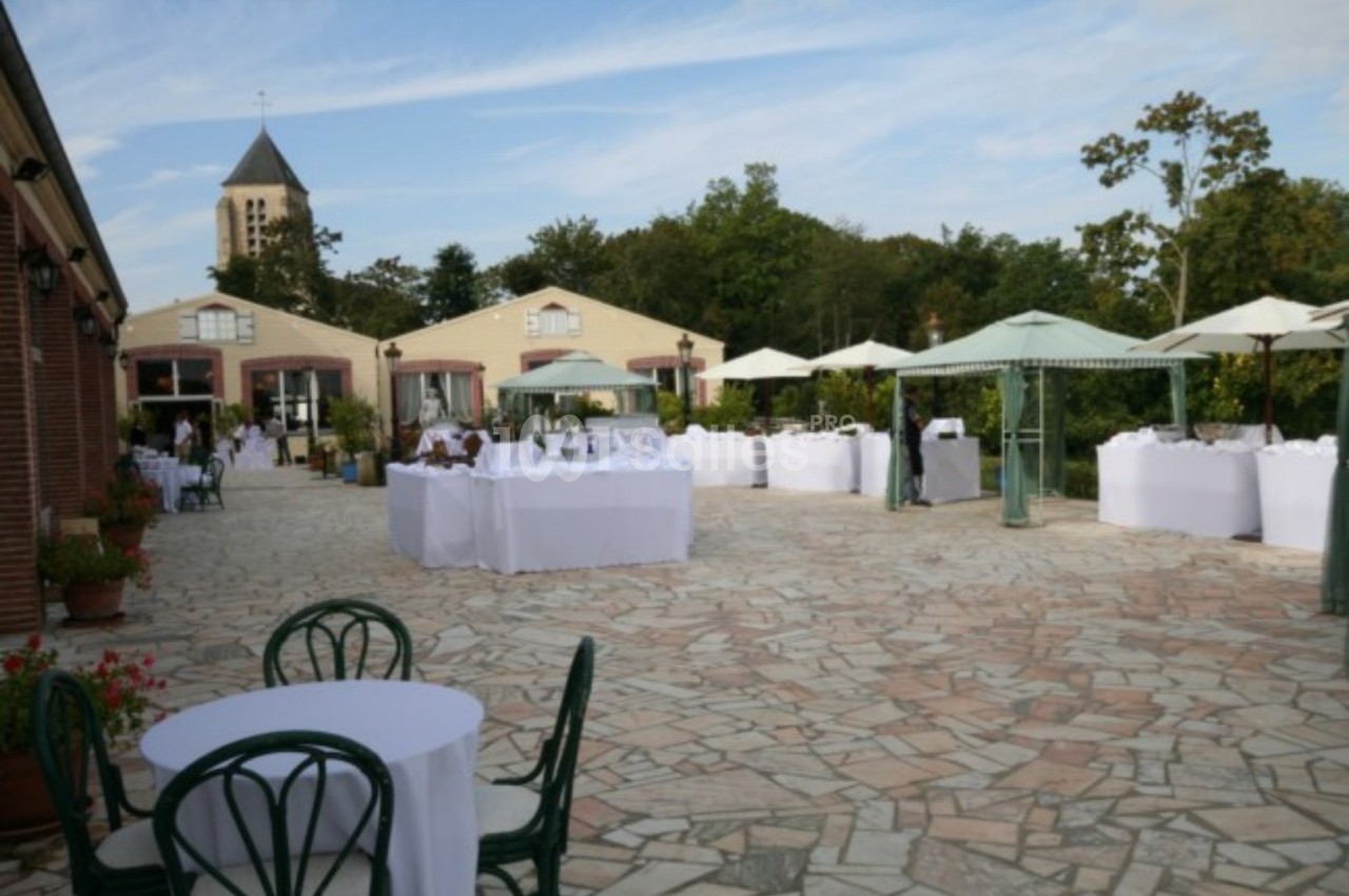 Terrasse pavée avec tables dressées, parasols et bâtiments en arrière-plan, entourée de verdure.