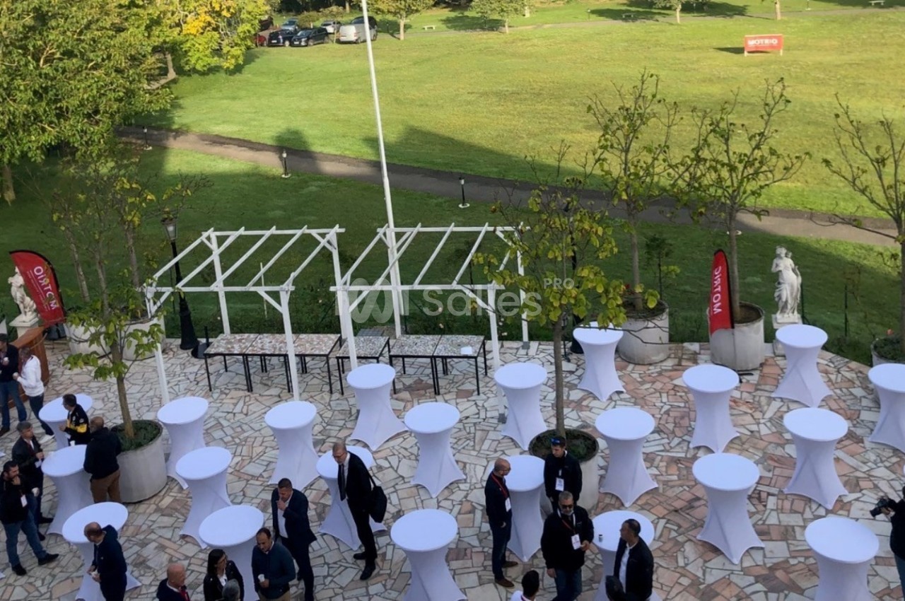 Vue aérienne d'une terrasse avec des tables hautes blanches, des invités debout et un jardin en arrière-plan.