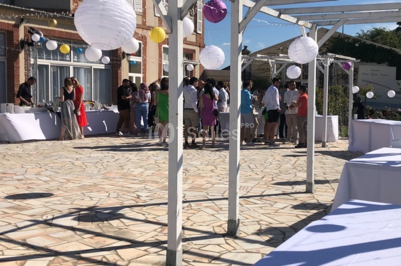Groupe de personnes rassemblées sur une terrasse ensoleillée décorée de lanternes blanches et de tables dressées.