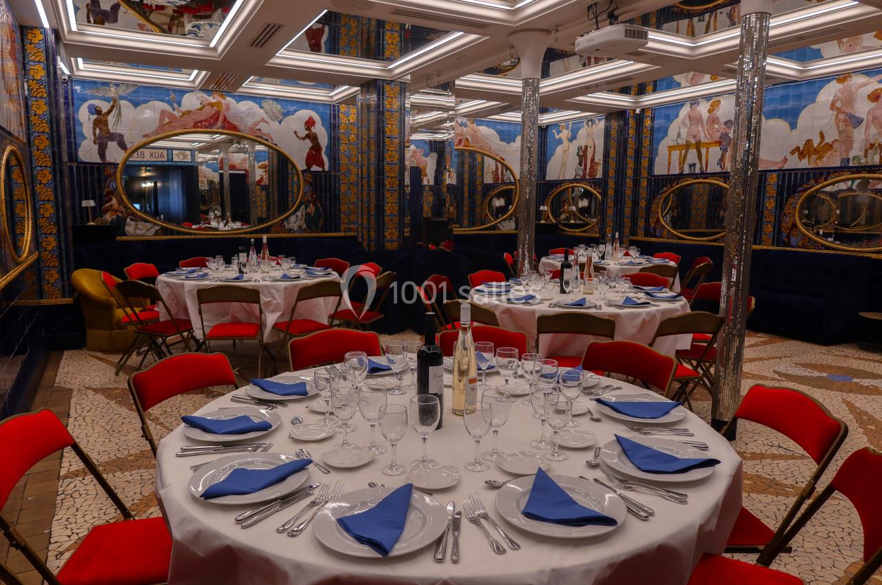 Salle de réception élégante avec tables rondes dressées, nappes blanches, serviettes bleues et miroirs ornés sur les murs.
