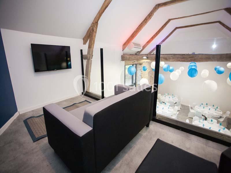 Location salle Plélan-le-Grand (Ille-et-Vilaine) - Domaine Le 5b #12 Vue d'une mezzanine avec canapé, télévision murale et aperçu d'une salle décorée de lanternes bleues et blanches.