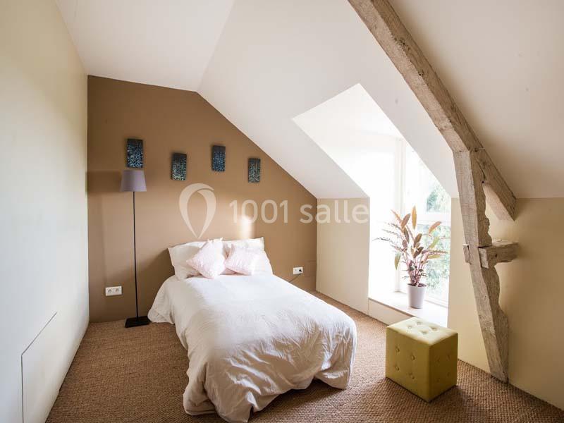 Location salle Plélan-le-Grand (Ille-et-Vilaine) - Domaine Le 5b #25 Chambre mansardée avec lit simple, mur beige, poutre apparente, lampe sur pied et fenêtre laissant entrer la lumière.