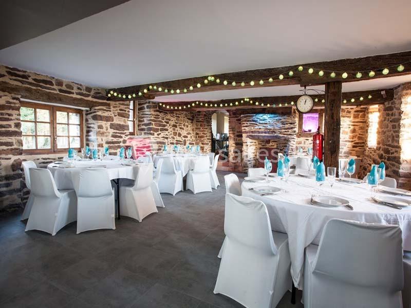 Location salle Plélan-le-Grand (Ille-et-Vilaine) - Domaine Le 5b #9 Salle en pierre avec tables rondes dressées de nappes blanches, chaises blanches et décorations lumineuses suspendues.