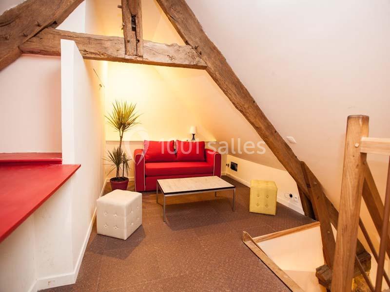 Location salle Plélan-le-Grand (Ille-et-Vilaine) - Domaine Le 5b #28 Salon mansardé avec poutres apparentes, canapé rouge, table basse, poufs et éclairage doux.